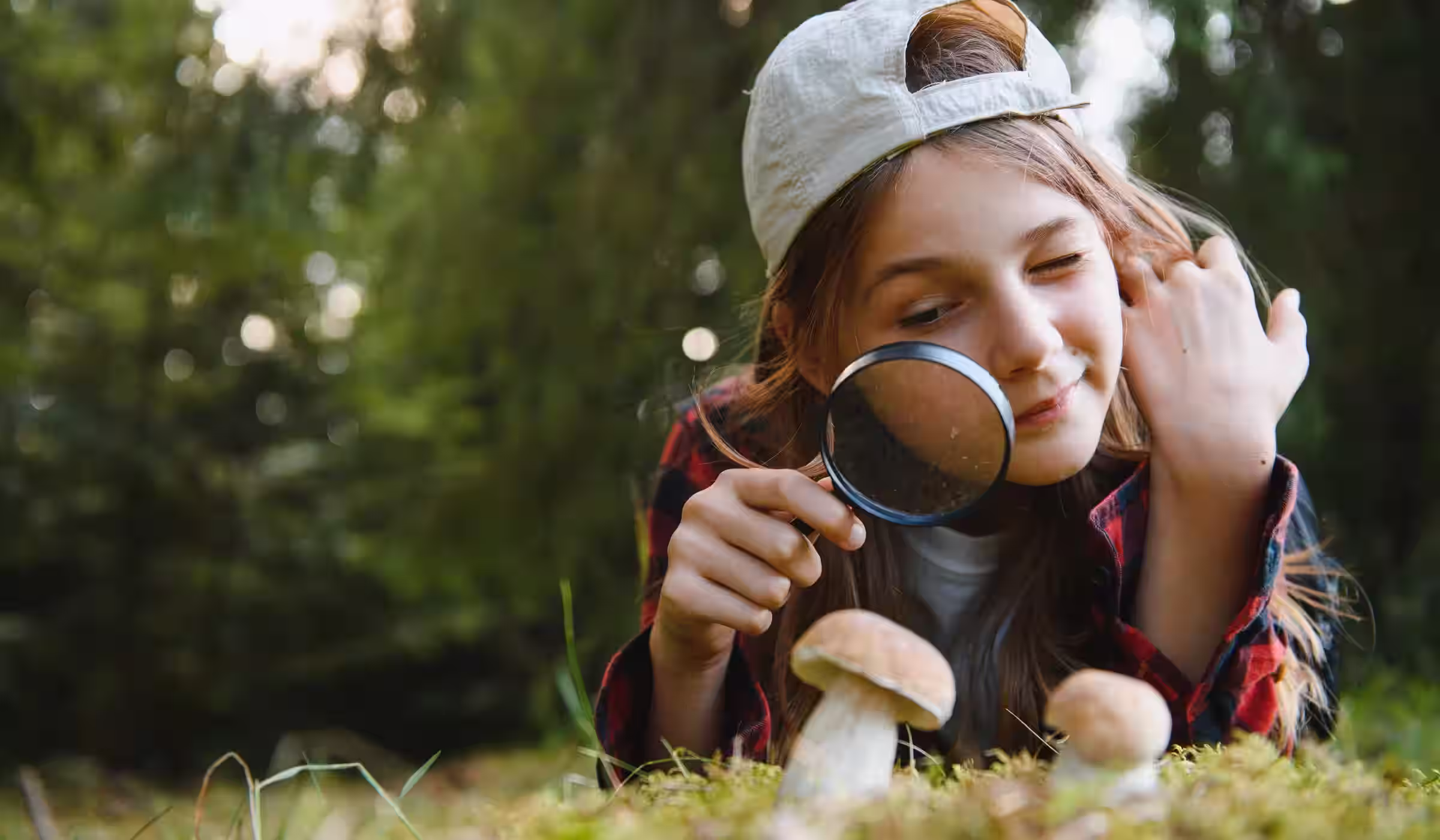 A child lies in the grass examining mushrooms through a magnifying glass.